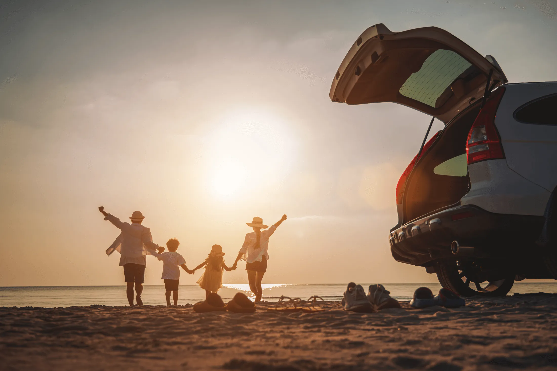 backs of a family of four all holding hands on a sunsetted beach with a car covered by Envela auto insurance in the foreground